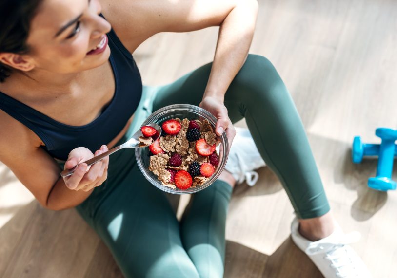 Athletic woman eating a healthy bowl of muesli with fruit sitting on floor in the kitchen at home